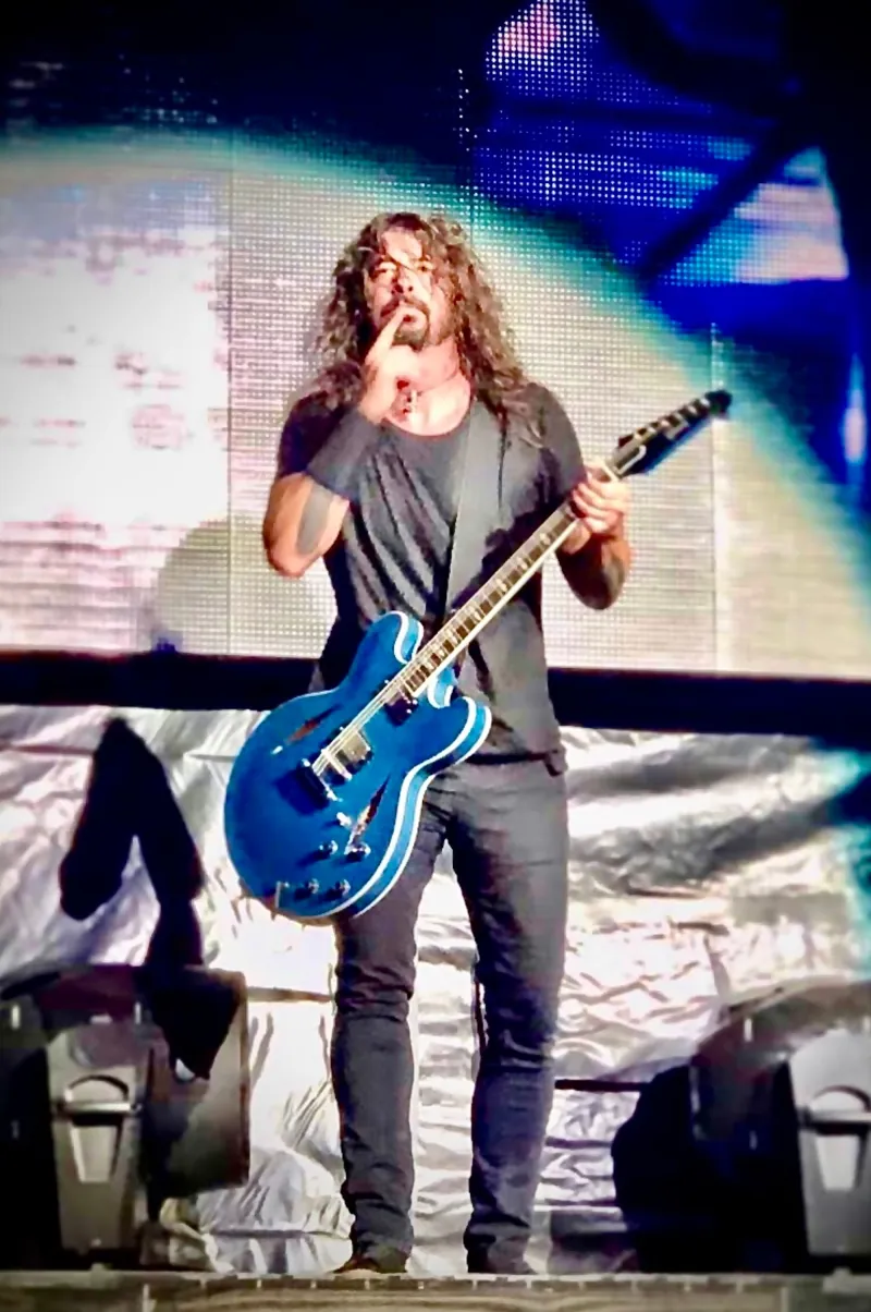 Dave Grohl with a blue semi-hollow guitar, finger raised to his lips, in front of a glowing LED wall