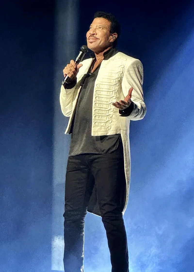 Lionel Richie singing under a blue spotlight in a white jacket