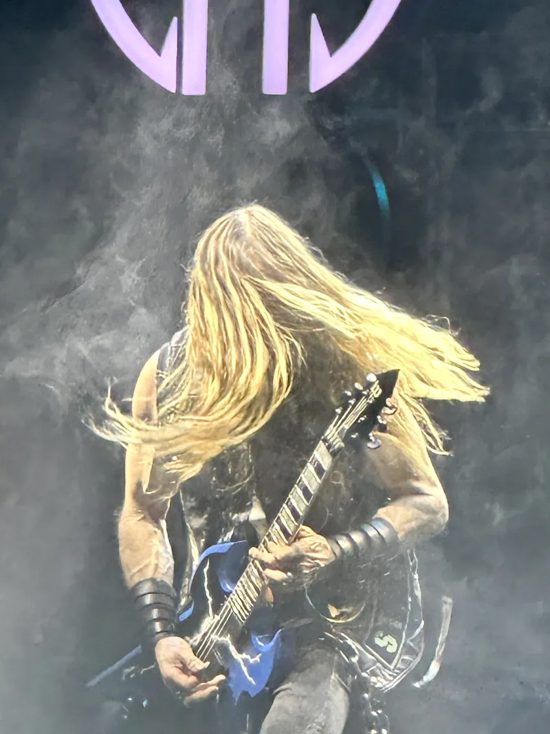 Zakk Wylde mid head-toss with long blond hair, playing guitar through stage smoke